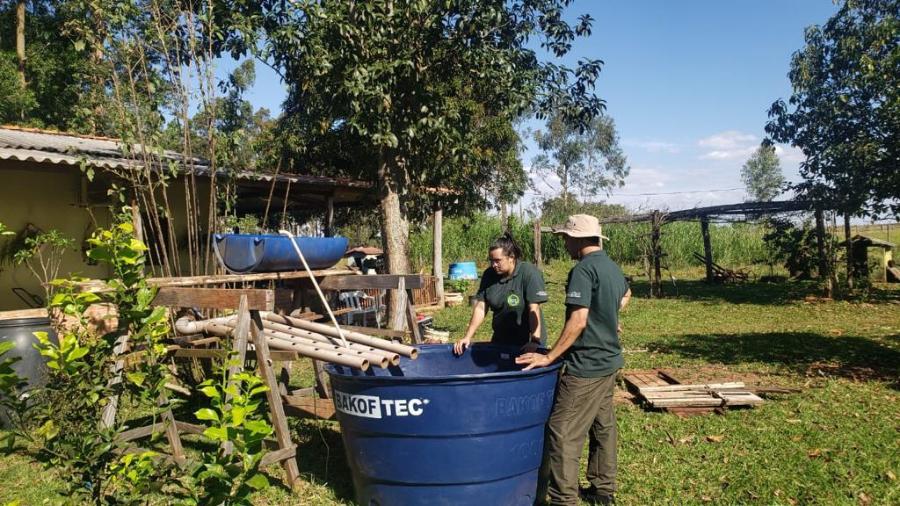 Fotografia de aluna da UFGD junto a um homem do assentamento, eles estão verificando o tanque de peixes que faz parte do sistema de aquaponia