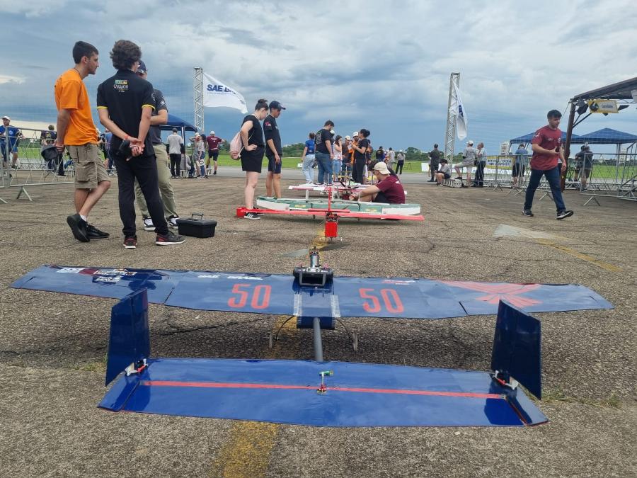 Na foto podemos ver em primeiro plano um aeromodelo azul, pousado em uma pista de vôo. Ao fundo da foto vemos cerca de oito jovens olhando outros aeromodelos. 