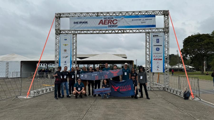 A foto ontém 15 jovens que estão posando em frente a um pórtico onde está escrito SAE Brasil AeroDesign