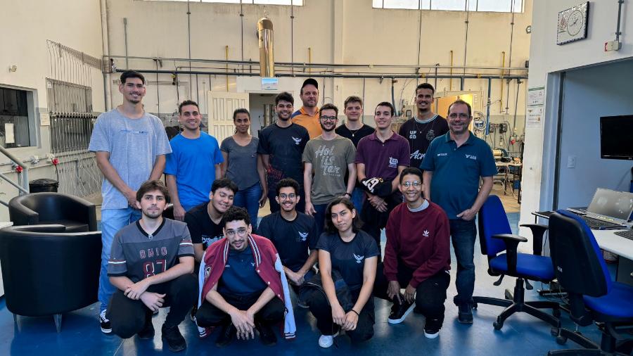Na foto 16 pessoas, a maioria jovens estudantes, estão posando em um laboratório de física, onde tem dois computadores e outros equipamentos. 