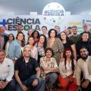 Foto com mais de 20 pessoas ao fundo um paine escrito Programa Mais Ciência na Escola